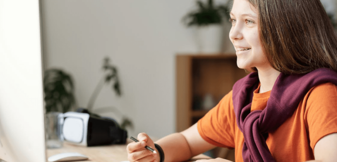 A girl wearing an orange shirt sitting at a computer desk while smiling at the PC screen