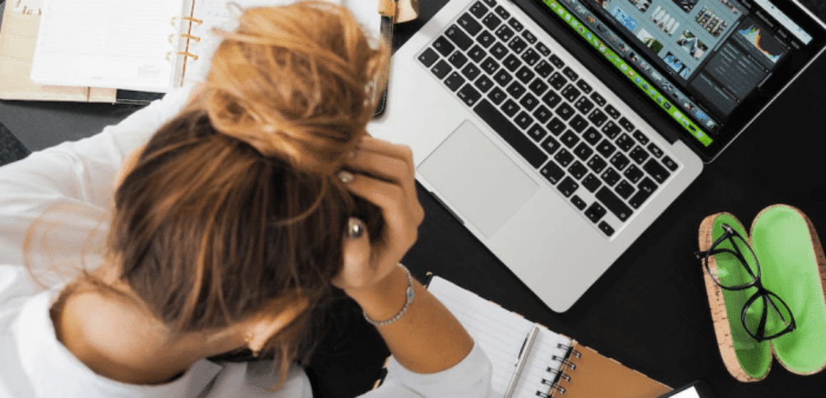 A woman is sitting in front of a MacBook with a notebook lying open as she holds her head in frustration